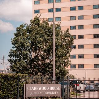 An exterior view of Clarewood House Senior Community, featuring a tall apartment building with a surrounding fence and trees.