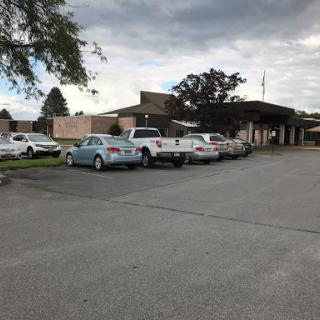 A view of a parking lot with multiple vehicles parked in front of a building under a cloudy sky.