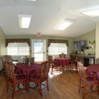 A well-lit dining area with multiple tables and chairs, featuring large windows and a wooden floor.