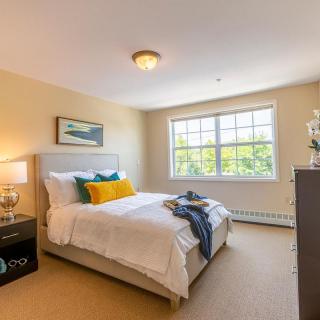 A cozy bedroom with a large window letting in natural light, featuring a neatly made bed with colorful pillows and simple decor.