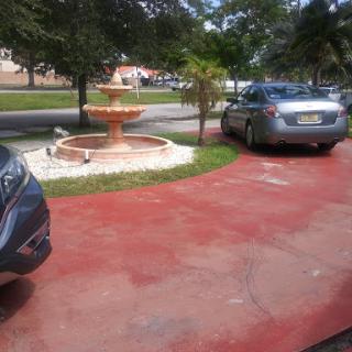 A sunny day featuring parked cars beside a decorative fountain surrounded by greenery.