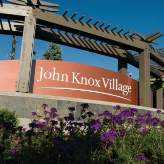 A sign for John Knox Village surrounded by purple flowers and outdoor structures.
