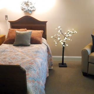 A cozy bedroom featuring a neatly made bed with decorative pillows, a soft armchair, and a unique floor lamp resembling a lit tree.