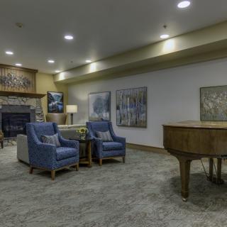 A cozy living room featuring armchairs, a grand piano, a fireplace, and decorative artwork.
