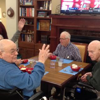 A group of seniors sitting around a table, engaging in conversation while watching TV.