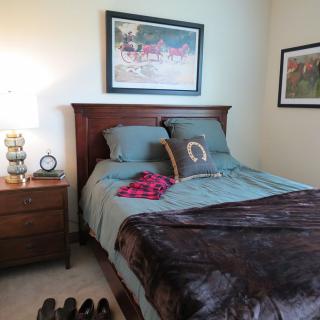 A cozy bedroom with a neatly made bed, featuring decorative pillows and a soft blanket. A nightstand with a lamp and alarm clock is on the side, and artwork adorns the walls.
