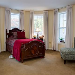 A cozy bedroom with a traditional wooden bed and red bedding, featuring bay windows with curtains.
