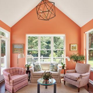 A cozy living room with orange walls, featuring a sofa and armchairs around a coffee table, with large windows overlooking a garden.