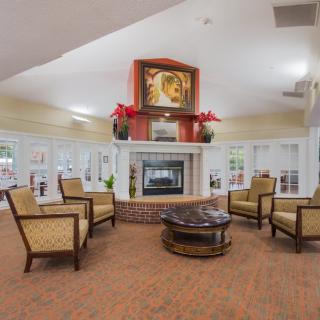 A cozy living room with a central fireplace, surrounded by comfortable chairs and a coffee table, featuring artwork and plants for decoration.