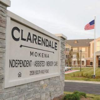 A sign for Clarendale featuring Independent, Assisted Living, and Memory Care services, with a building and an American flag in the background.