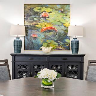 A stylish dining room with a vibrant painting, featuring koi fish and lily pads, flanked by two decorative lamps on a sideboard.