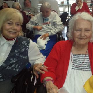 Two elderly women smiling and sitting in wheelchairs indoors.