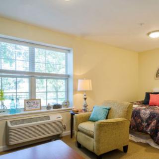 A cozy living space with a large window, comfortable chair, and a neatly made bed.