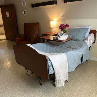 A cozy hospital patient room featuring a bed with a blue cover, a comfortable chair, and a bedside lamp illuminating the space.