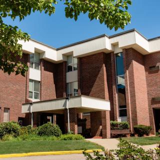 A modern brick building with a welcoming entrance, surrounded by greenery under a clear blue sky.