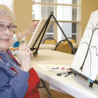 A senior woman participates in an art class, working on a painting of a tree on canvas.