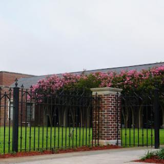 A view of a brick building surrounded by a wrought iron fence and flowering plants.