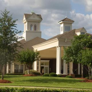 A large building with two small towers and white columns, surrounded by trees and landscaping under a cloudy sky.