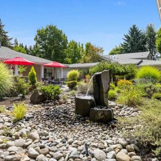 A well-maintained garden with a central fountain, surrounded by rocks and vegetation, featuring patio furniture with red umbrellas.