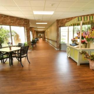 A bright hallway with wooden floors, tables, chairs, and decorative flowers along the side.