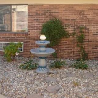 A small garden area featuring a tiered fountain, surrounded by gravel and plants, set against a brick wall.