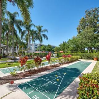 A sunny outdoor scene featuring a shuffleboard court surrounded by lush tropical gardens and palm trees.