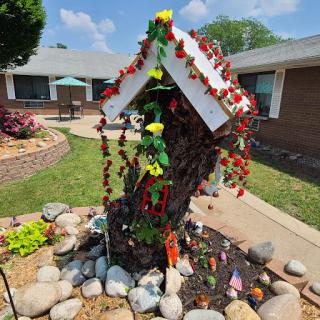 A colorful garden decoration featuring a tree adorned with artificial flowers and surrounded by stones.