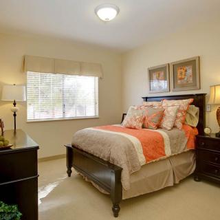 A cozy bedroom with a neatly made bed featuring orange and beige bedding, flanked by bedside tables with lamps, and artwork hanging above the bed.