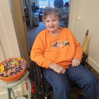 A smiling woman in a wheelchair, dressed in an orange sweater for Halloween, sitting beside a bowl of candy.