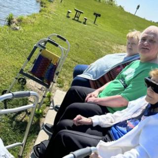 A group of people enjoying a sunny day at the park, seated near the water with a walker nearby.