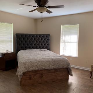 A minimalist bedroom with a large bed, nightstands, and a ceiling fan, illuminated by natural light from two windows.