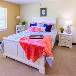 A cozy bedroom with a neatly made bed, colorful pillows, and a bright coral blanket. The room features a nightstand with a lamp, plants for decoration, and natural light coming through the window.