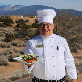 A chef proudly presents a beautifully plated dish against a scenic backdrop.