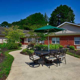 A sunny outdoor patio with a table, chairs, and an umbrella surrounded by a landscaped garden.