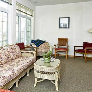 A cozy living room featuring wicker furniture and large windows, with a carpeted floor and indoor plants.