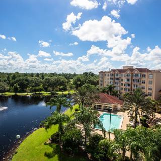 A scenic view of a resort with a pool, surrounded by palm trees and overlooking a tranquil lake under a bright, cloudy sky.