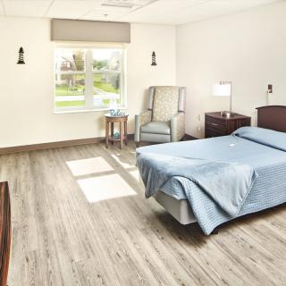 A clean and minimalist hospital room with a single bed, a chair, and a window letting in natural light.