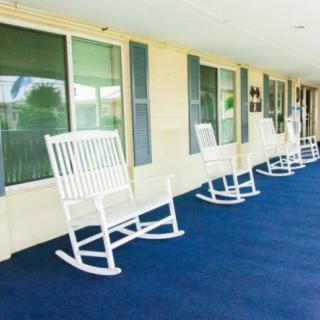 A row of white rocking chairs lined up on a porch with blue carpeting.