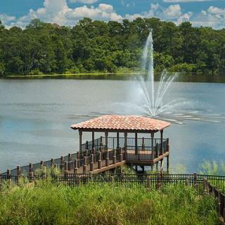 A scenic view of a gazebo by a lake with a fountain and lush greenery.