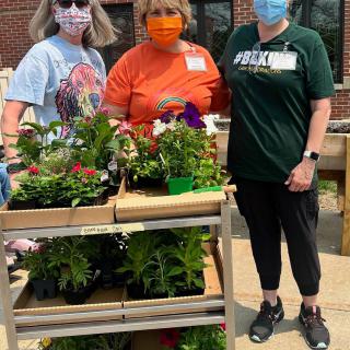 Three people wearing masks stand beside a trolley filled with plants and flowers.