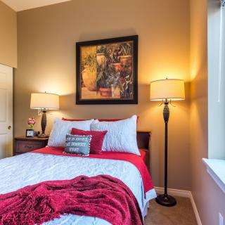 A cozy bedroom with a neatly made bed, featuring red and white bedding, flanked by two floor lamps and adorned with wall art.