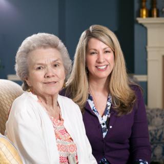 An elderly woman and a younger woman smiling together in a living room.