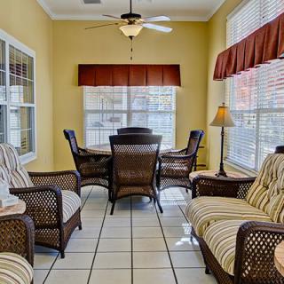 A cozy sunroom featuring rattan furniture and a dining table, illuminated by natural light streaming through large windows.