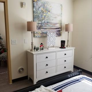 A cozy bedroom with a white dresser, adorned with table lamps and decorative items, and a serene landscape painting on the wall.