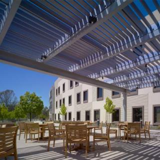 A sunny outdoor patio area with wooden chairs and tables under a modern pergola, adjacent to a contemporary building.