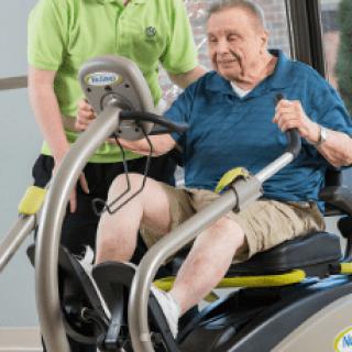 An elderly person exercising on a recumbent bike with assistance.