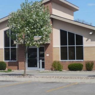 A commercial building with large windows and a tree in front, located next to an empty parking lot.