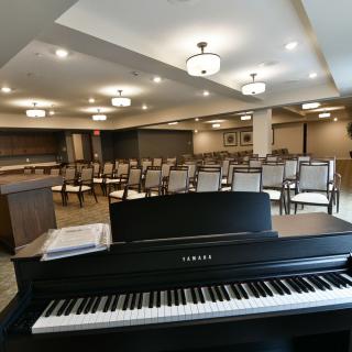An empty auditorium with a piano in the foreground and rows of chairs facing a stage.