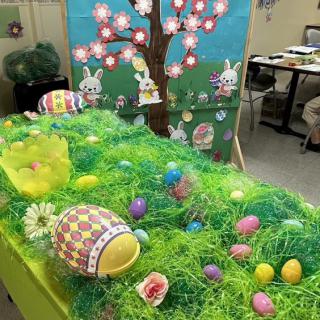 A festive Easter table display featuring colorful eggs, a grassy setup, and bunny decorations with a painted backdrop.