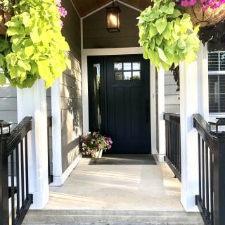 A welcoming front porch with hanging and potted plants, leading to a dark-colored door.
